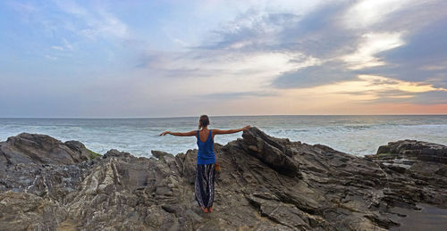 Rear view of woman with arms outstretched standing on rock at beach against sky