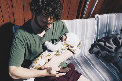 Father with baby boy using mobile phone while sitting on chair during sunny day