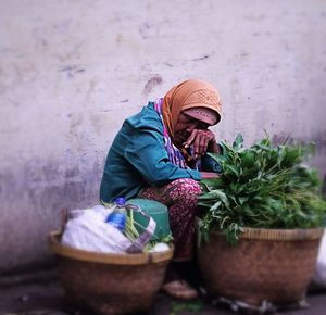Senior vendor sleeping while selling leaf vegetables against wall