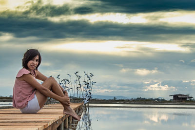 Portrait of mid adult woman sitting on pier over lake against cloudy sky during sunset