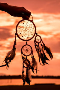 Cropped hand holding dreamcatcher against orange sky