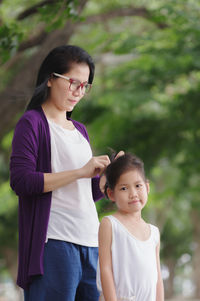 Mother tying daughter hair while standing against tree