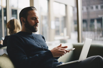 Businessman holding smartphone while usign laptop at office lobby