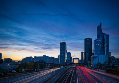 Light trails on road amidst buildings against sky at sunset