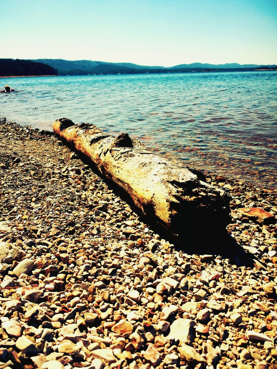 water, beach, clear sky, sea, tranquility, tranquil scene, shore, scenics, nature, beauty in nature, stone - object, pebble, sky, rock - object, sand, idyllic, surface level, outdoors, blue, sunlight