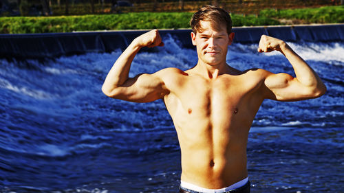 Portrait of shirtless man flexing muscles against river