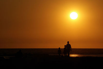 Silhouette people sitting on beach against orange sky