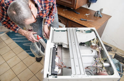 Middleaged master in checkered suit unscrews the lid of  washing machine with screwdriver for repair