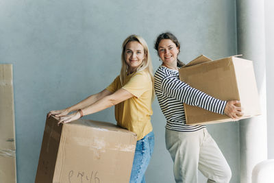 Happy adult women relocating house lifting cardboard box with life stuffs. friend helping.