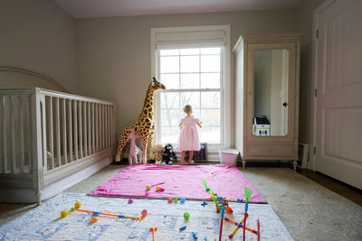 Toddler girl looks out the window of her nursery, toys on the floor