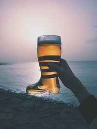 Cropped hand of woman holding boot shape beer glass at beach against sky during sunset