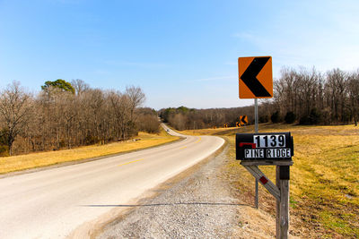 Mailbox and arrow signs on field by empty road against clear sky