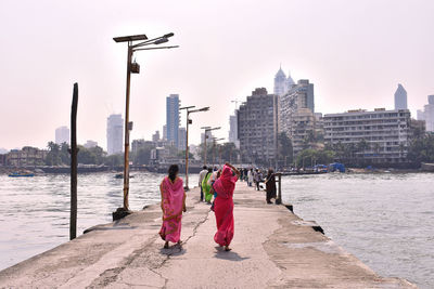 Rear view of people walking on footpath by buildings against sky