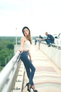 Young woman leaning on railing while standing on bridge against clear sky