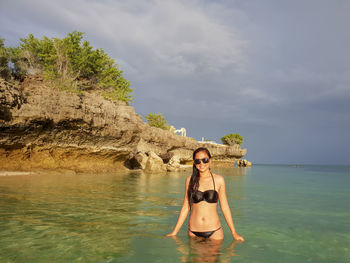 Portrait of young woman in bikini standing by sea against sky