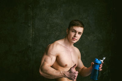 Portrait of shirtless man with bottle gesturing thumbs up against wall