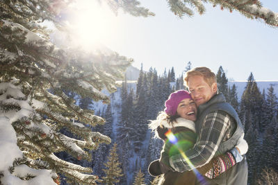Couple embracing while standing in snowy forest against clear sky