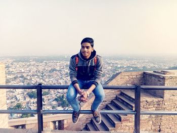 Full length of young man sitting on railing against cityscape