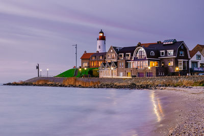 Illuminated buildings by sea against sky at dusk