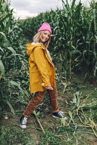Stylish teenage girl in a yellow raincoat and a hot pink cap is dancing and laughing on a cornfield