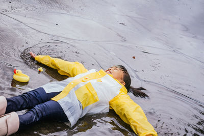 High angle of content asian kid in rubber boots and slicker lying in rippled puddle on rainy day