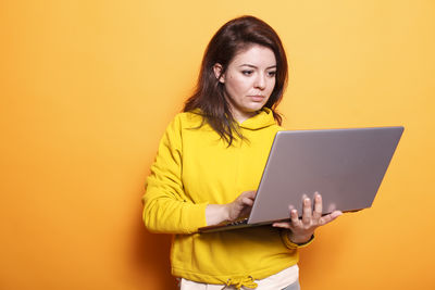 Portrait of young woman using laptop while sitting against yellow background