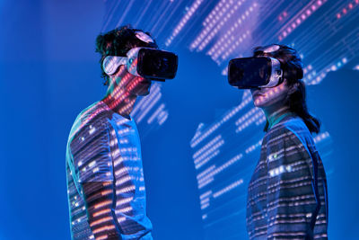 Side view of couple in modern vr goggles while standing on blue background with glowing lights in studio