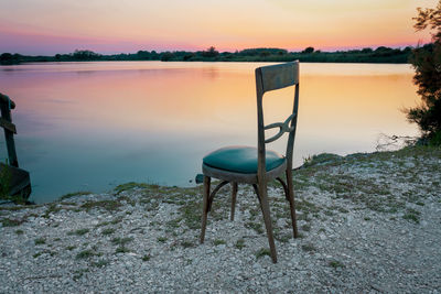 Chair by lake against sky during sunset