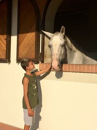 Side view of boy stroking horse peeking through window in stable