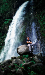 Scenic view of waterfall with man sitting on rock in forest