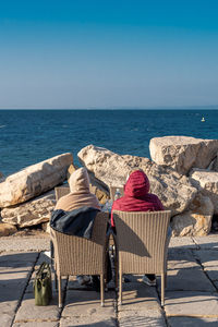 Rear view of woman sitting by sea against clear blue sky