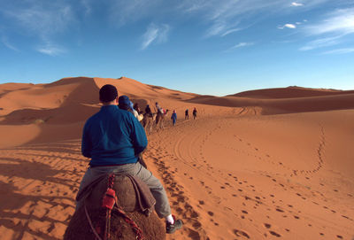 Rear view of people riding camels at desert against sky