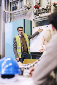 Mature male worker discussing plan on whiteboard to colleagues in factory