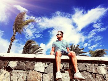 Low angle view of young man sitting on retaining wall against sky during sunny day