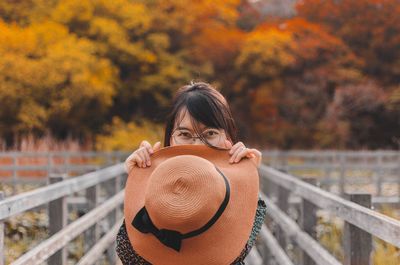 Portrait of young woman holding hat against trees on footbridge during autumn