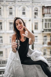 Portrait of cheerful woman having red wine while wrapped in blanket at balcony