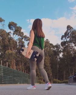 Rear view full length of young woman holding skateboard walking on road