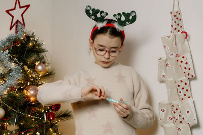 A girl opens a candy from an advent calendar while standing near a christmas tree.