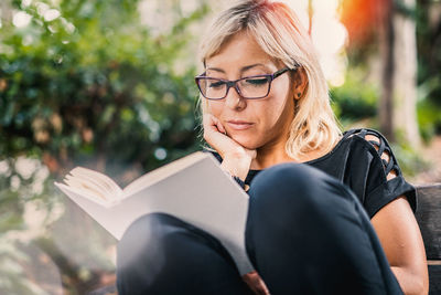 Young woman reading book while sitting on bench in park