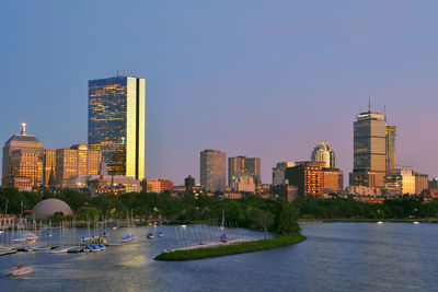 Modern buildings by river in city against clear sky during sunset