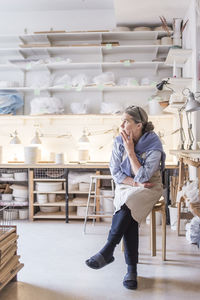 Senior female potter looking away while sitting on stool against shelves in workshop