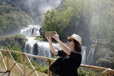 Smiling woman taking selfie while standing by railing with waterfall in background