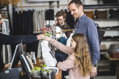 Side view of daughter receiving shopping bag from cashier by father at checkout counter in clothing store