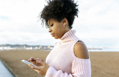 Beautiful woman with curly hair using mobile phone while sitting against clear sky