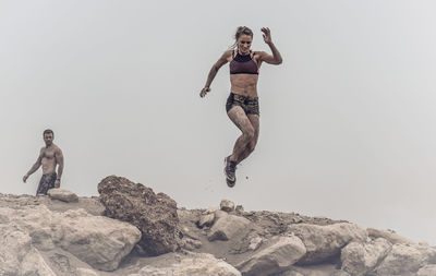 Man standing while woman jumping at dirt road against clear sky