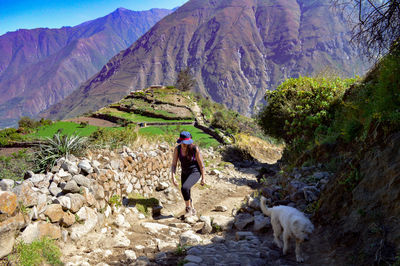 Full length of woman hiking with dog on rocky footpath against mountain