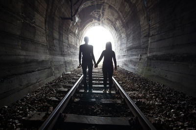 Rear view of female friends holding hands while standing on railroad track in tunnel