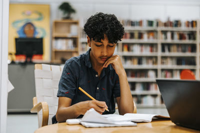 Focused teenage boy studying with hand on chin while sitting near table in library at junior high school