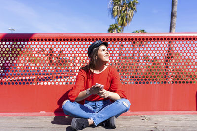 Teenage girl using mobile phone while wearing red top in city against railing during sunny day