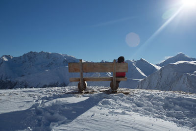 Rear view of woman sitting on bench at snow covered field against clear sky